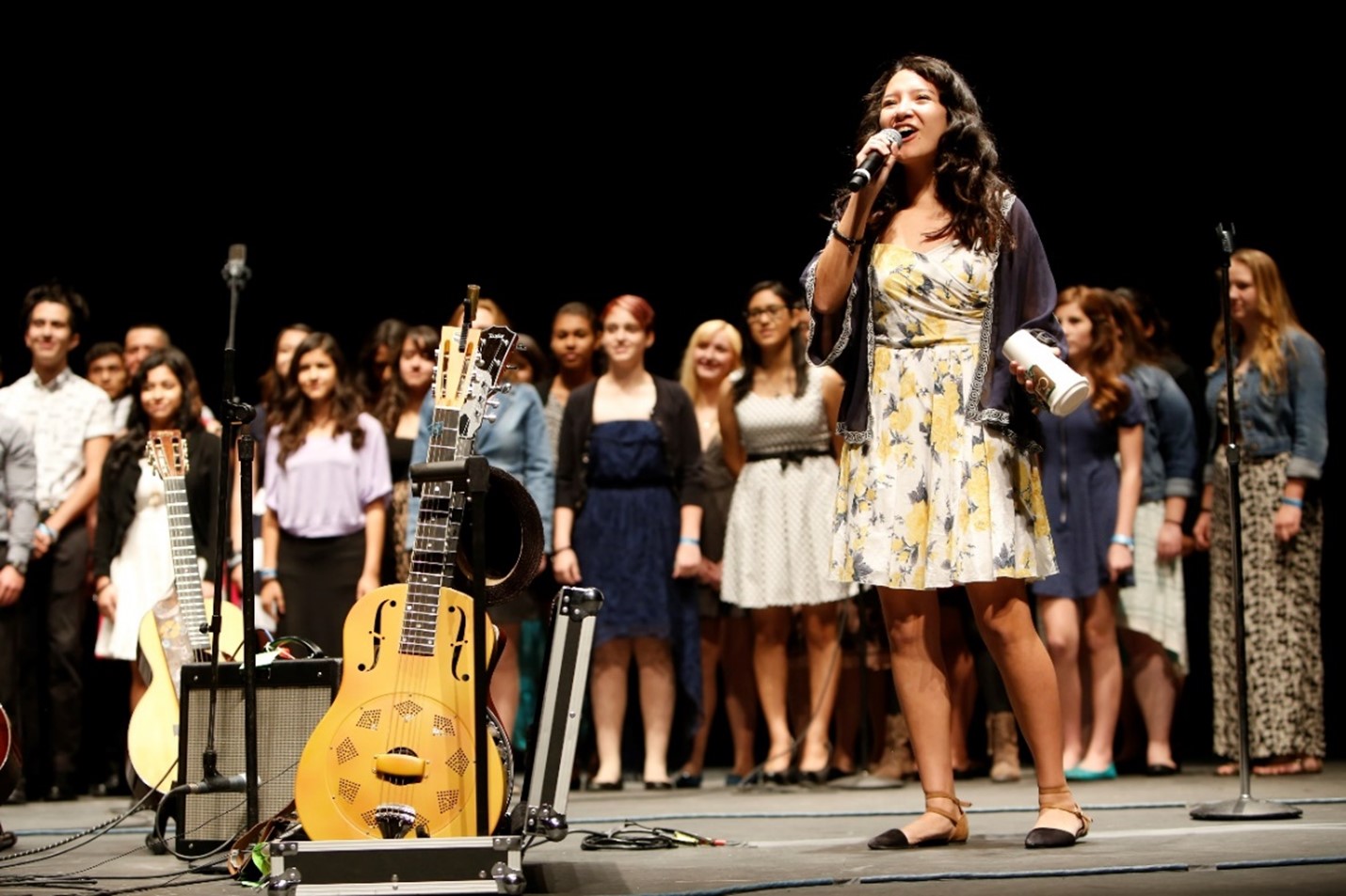 student on stage with microphone appears to be singing. There is a group of students behind her on the stage and two acoustic guitars to the left of the student.